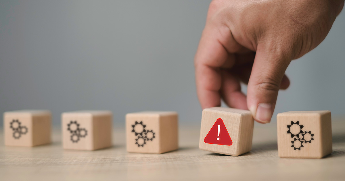 Eine Hand, die einen Holzblock mit einem Warnsymbol aus einem Satz von Blöcken herausgreift, was die Identifizierung von Problemen, das Risikomanagement und die Problemerkennung im Prozess symbolisiert.