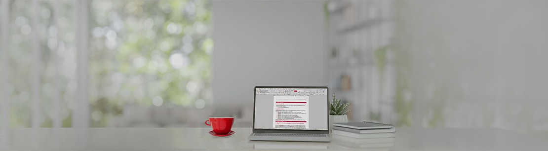 Einen Laptop auf einem Tisch. Auf dem Bildschirm ist ein Word-Dokument geöffnet. Daneben stehen eine rote Tasse, eine kleine Pflanze und ein paar Bücher.