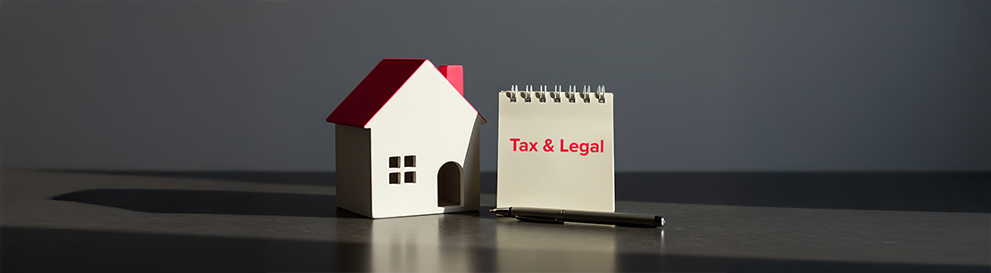 Ein kleines Modellhaus, einen Notizblock mit der Aufschrift „Tax & Legal“ und einen Stift. Alles steht auf einem Tisch.
